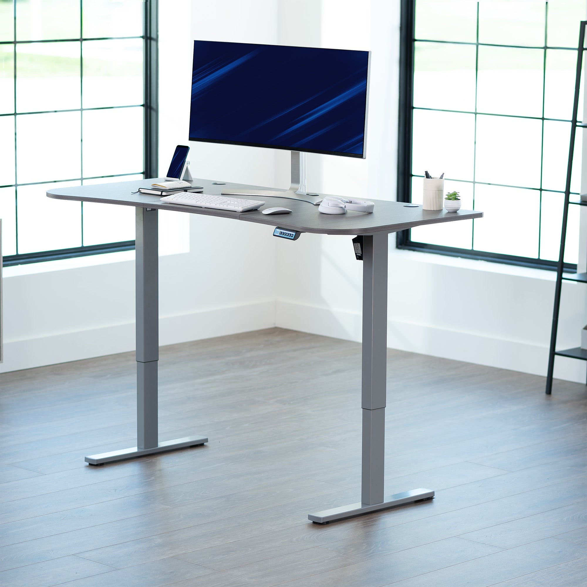 Dark grey top grey frame, electric, sit to stand, height adjustable desk in office setting.