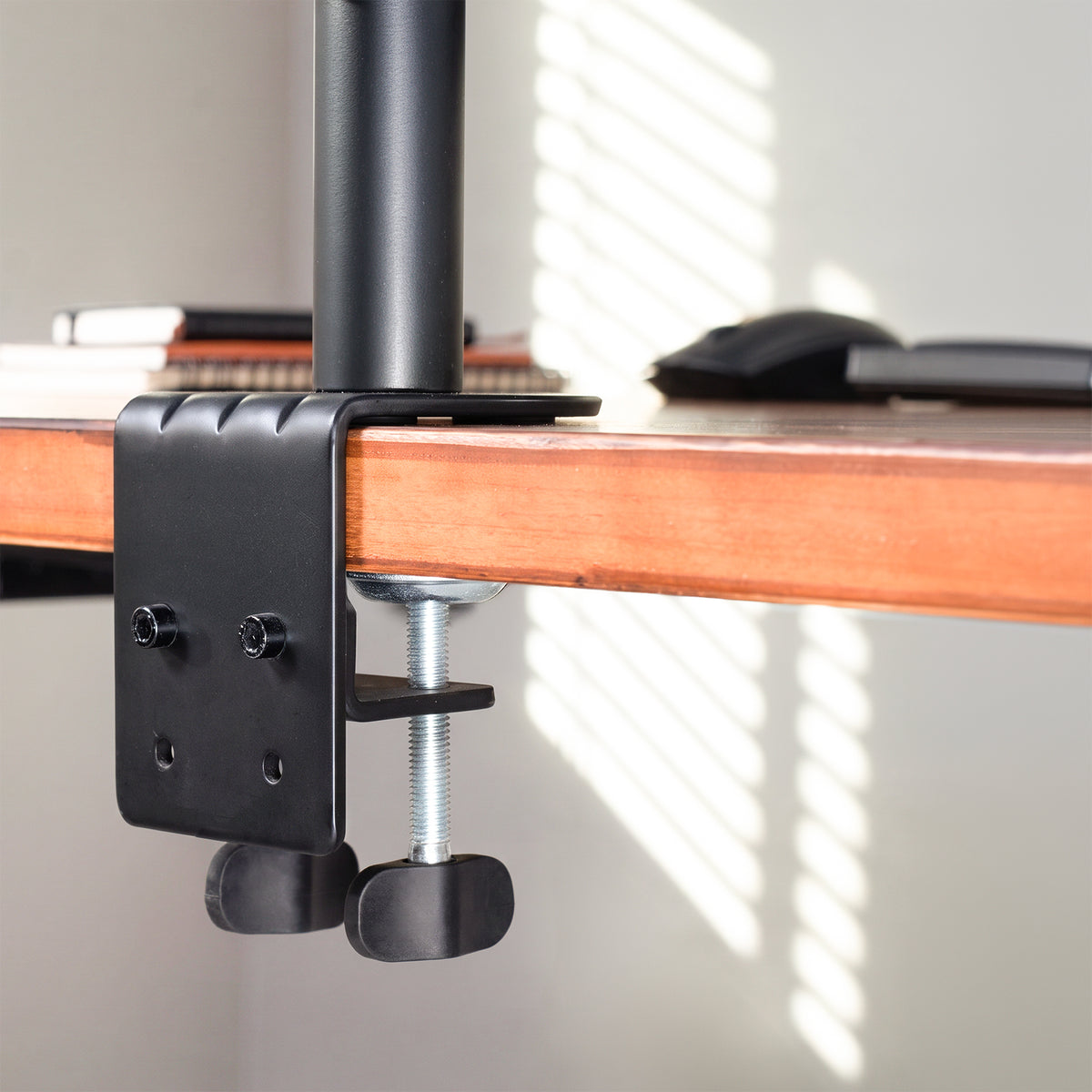 A close-up of a black desk clamp mount with a silver threaded screw and black rubber feet, attached to a wooden desk under a black desk pad. The background shows a softly blurred indoor setting with window blinds casting light shadows.