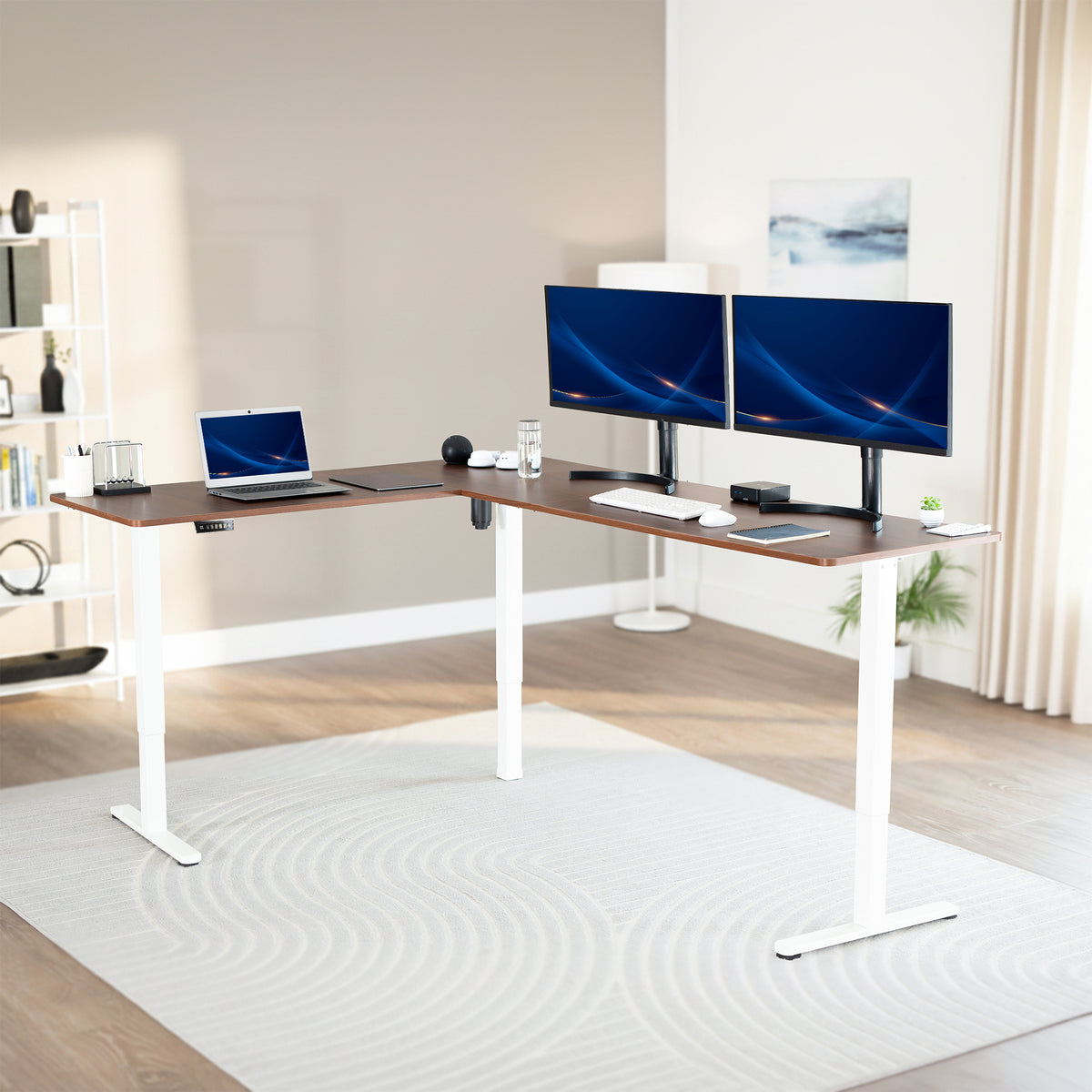 Modern office setup with a standing l-shaped desk, laptop, and two monitors in a bright room.