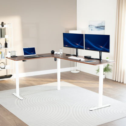 Modern office setup with a standing l-shaped desk, laptop, and two monitors in a bright room.