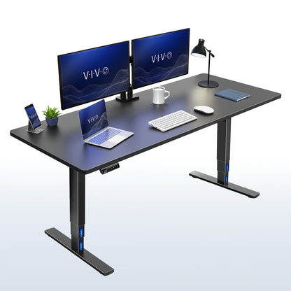 A black modern electric height-adjustable desk with a sleek metal T-style base. The desk is styled with a dual-monitor setup, a laptop, a smartphone on a charging stand, a small potted plant, a white mug, a keyboard, a mouse, and a desk lamp.