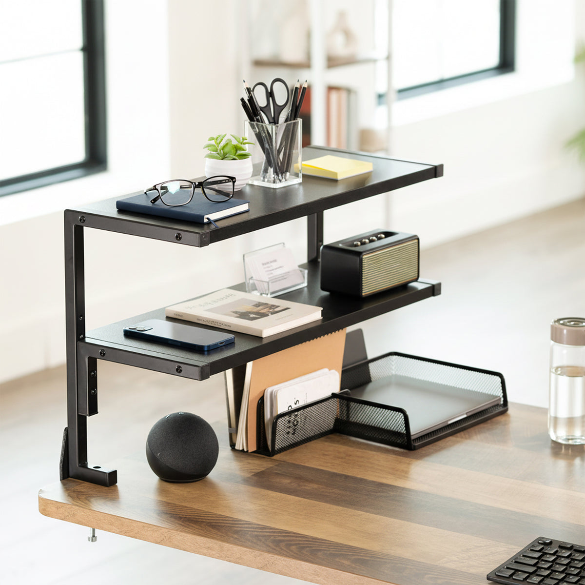 A black metal three-tier desktop organizer mounted on a wooden desk in a bright, modern home office. The shelves are styled with various office accessories including a cup of pens, glasses, a small plant, a speaker, a notebook, and a desk tray.