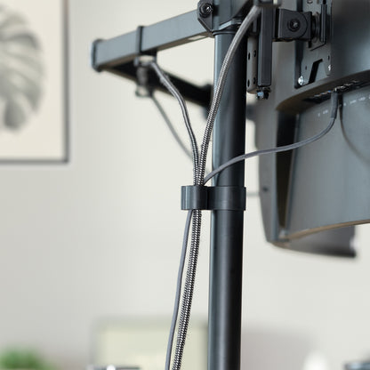 Close-up of a black monitor stand with cable management and mounting hardware, shown in a softly blurred indoor setting.